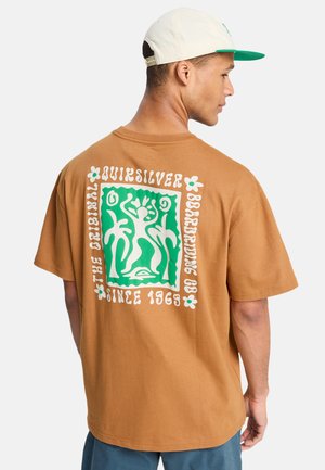 Junger Mann trägt ein beige Quiksilver T-Shirt mit grün-weißer Grafik einer tanzenden Figur und Palmen, kombiniert mit einer weißen und grünen Mütze.