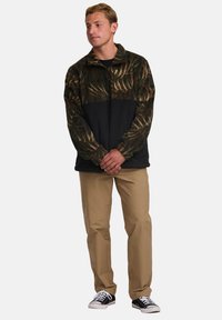 Fleece pullover with a green and brown leaf pattern on top, black lower section, zippered collar, and long sleeves, paired with khaki pants.