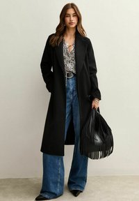 Lungo cappotto nero in lana, abbinato a una maglietta a fantasia, jeans larghi blu e una borsa a tracolla nera con frange. Scarpe nere a punta completano il look.