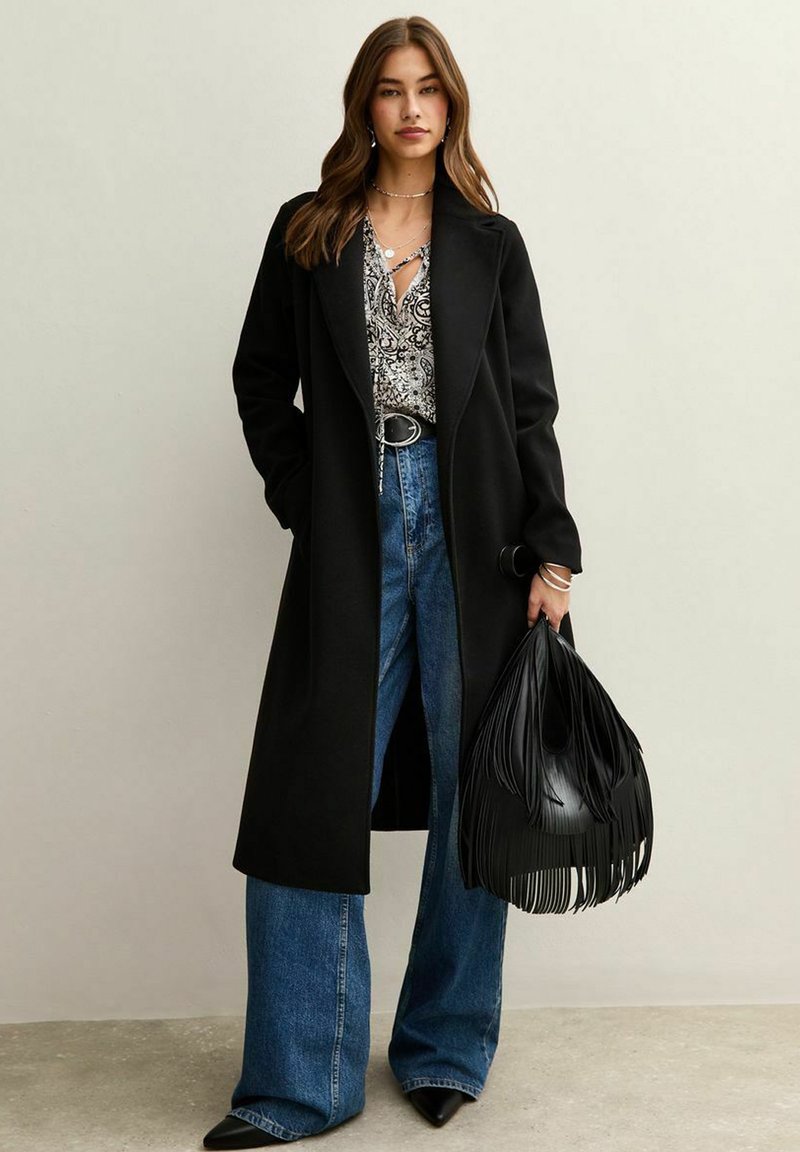 Lungo cappotto nero in lana, abbinato a una maglietta a fantasia, jeans larghi blu e una borsa a tracolla nera con frange. Scarpe nere a punta completano il look.