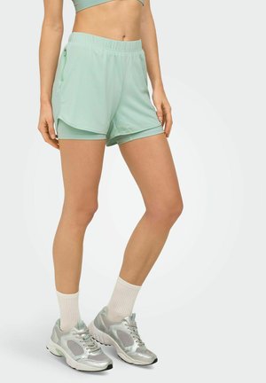 Mujer con pantalones cortos deportivos en capas de color verde claro, sujetador deportivo a juego, calcetines blancos hasta la pantorrilla y zapatillas de correr plateadas, de pie con las piernas cruzadas.