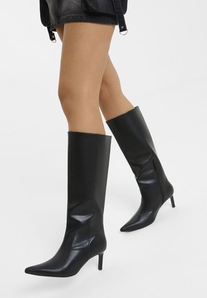 Jambes portant des bottes en cuir noires montantes à bout pointu avec des talons fins et une jupe en cuir noire sur un fond uni.