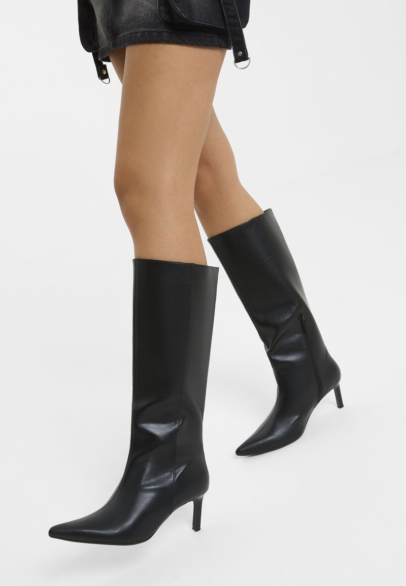 Jambes portant des bottes en cuir noires montantes à bout pointu avec des talons fins et une jupe en cuir noire sur un fond uni.