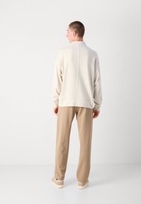 Helles beiges Sweatshirt mit hohem Kragen und einer Nahtdetails, kombiniert mit hellbraunen Hosen und hellen Schuhen, von hinten vor einem hellen Hintergrund betrachtet.