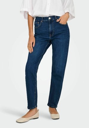 Femme portant un jean droit bleu foncé, une blouse blanche et des ballerines beige, debout avec une main dans la poche et un bras détendu.