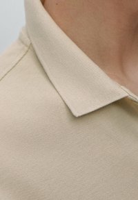 Beige texturerad polo skjorta med en vikbar krage, som har en slät tygfinish och subtila ribbor på kragkanterna.