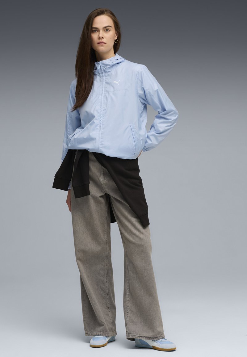 Veste à zip bleu clair avec capuche, arborant un logo. Portée sur un haut noir, associée à un jean gris ample et des baskets bleu clair.