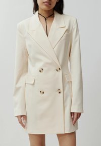 Cremefarbener, doppelt geknöpfter Blazer aus glattem Stoff mit goldenen Knöpfen, einem Reverskragen und zwei aufgesetzten Taschen.