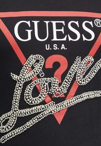 Camiseta de algodón negra con un gran logo triangular rojo que incluye el texto "GUESS U.S.A." y un script decorativo de brillantes plateados que dice "LOVE".