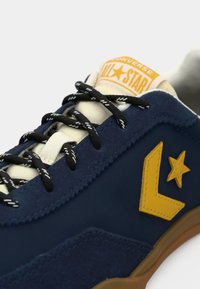 Blå ruskinds-sneakers med sort og hvide snørebånd, gult logo og orange "All Star" label på tungen; gummisål med lysebrune detaljer.