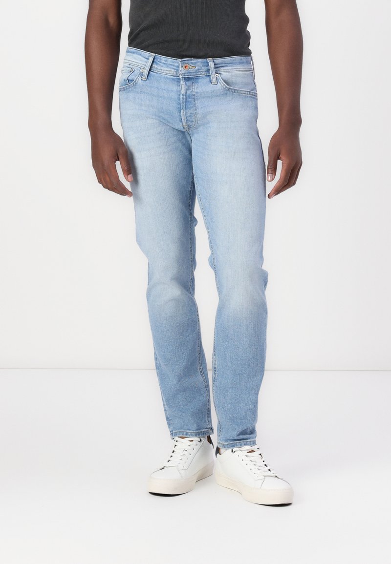 Jeans de denim azul claro con un corte ajustado, que cuentan con un diseño clásico de cinco bolsillos y un ligero desgaste. Llevados con zapatillas blancas y una camiseta sin mangas negra.