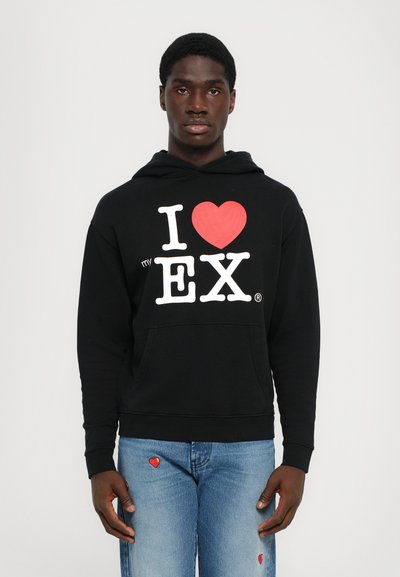Felpa nera in cotone con tasca frontale. Presenta scritte bianche e rosse, "I ❤️ my EX," e un design semplice. Indossata con jeans blu.