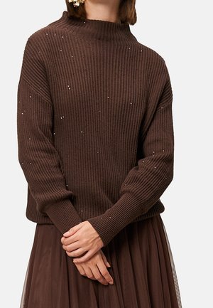 Kobieta w brązowym, prążkowanym swetrze z małymi białymi kropkami oraz w pasującej plisowanej brązowej spódnicy, z delikatnie splecionymi dłońmi z przodu.