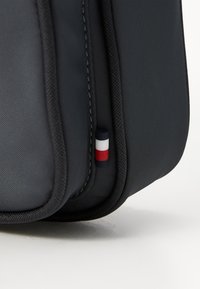 Borsa in tessuto grigia con una superficie liscia, caratterizzata da un piccolo logo tricolore (blu, bianco, rosso) su un lato e rifiniture nere lungo i bordi.