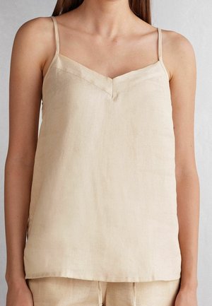Top smanicato beige in lino con sottili spalline e scollo a V indossato da una persona in piedi davanti a uno sfondo semplice.