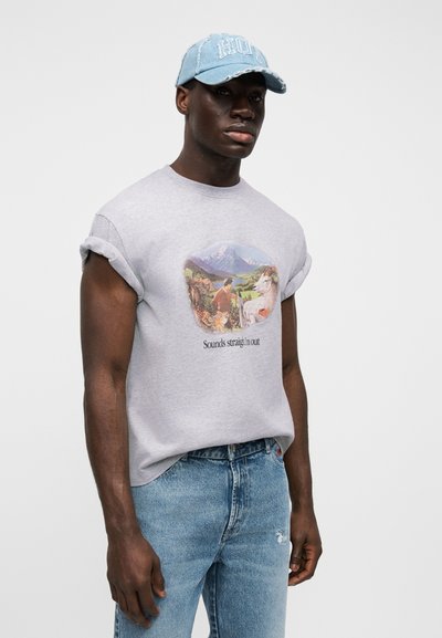 Sivé tričko s krátkym rukávom s farebnou grafikou zobrazujúcou krajinu a text, doplnené svetlomodrými rifľami a opotrebovanou šiltovkou.