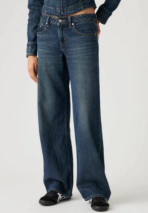 Wide-leg jeans in dark blue denim, featuring five-pocket design, logo hardware, and faded texture. Paired with black athletic shoes.