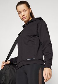 Hoodie zippé noir en tissu doux, comportant une capuche, des poches latérales et un accent bleu clair sur l'ourlet. Le mannequin tient un sac de sport noir.