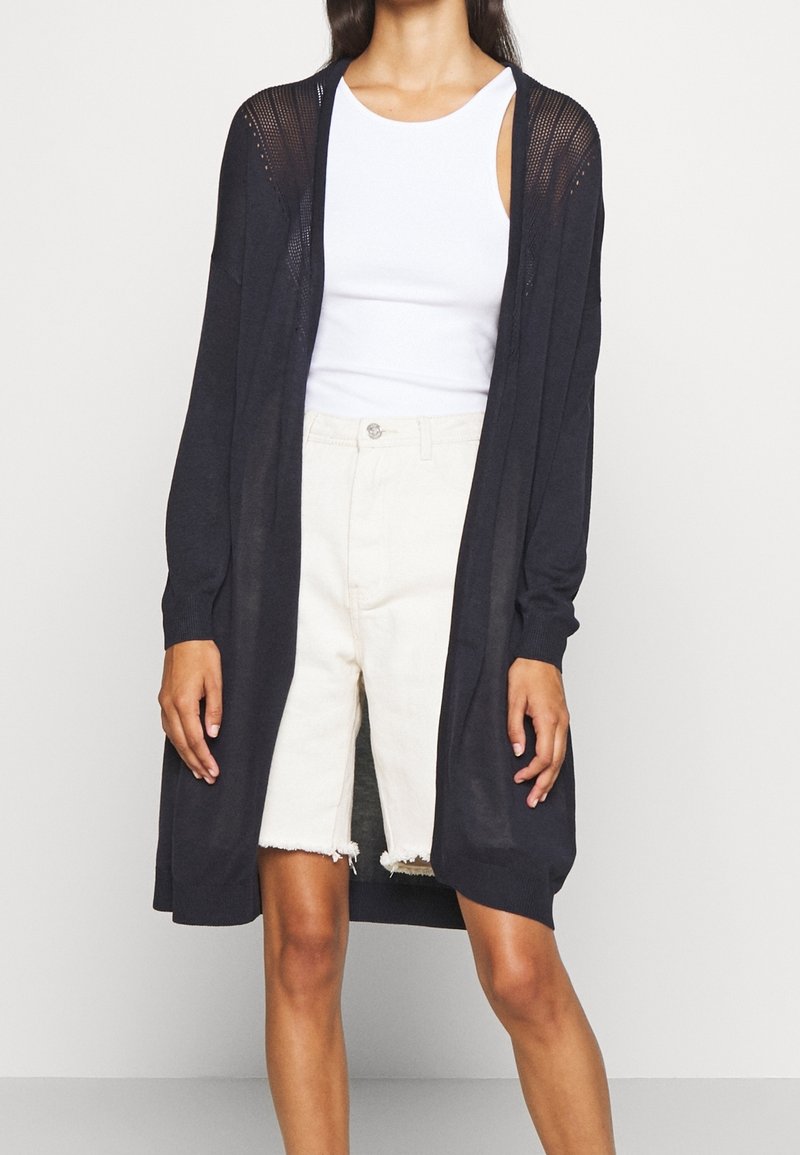 Femme portant un long cardigan noir ouvert devant, un top blanc sans manches et un short taille haute écru avec des bords effilochés.