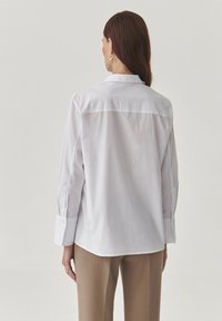 Camicia bianca a maniche lunghe con yoke posteriore e orlo curvo, realizzata in tessuto liscio. Abbinata a pantaloni marrone chiaro.