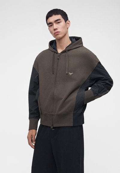 Jeune homme portant un sweat à capuche zippé bicolore marron et noir avec un petit emblème sur la poitrine, debout avec une main dans la poche.