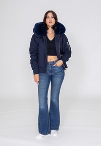 Marineblaues Bomberjacke mit großem Kunstfellkragen, Reißverschluss vorne, gerippten Bündchen und aufgesetzter Tasche. Kombiniert mit weit geschnittenen Jeans und weißen Sneakers.