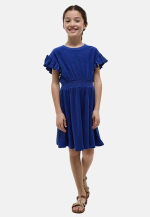 Fille souriante debout, portant une robe bleu roi à manches courtes avec des volants aux manches et des sandales tressées beige.