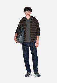 Giacca a vento nera con cappuccio, polo grigia, jeans scuri e scarpe da ginnastica grigie. Tiene uno zaino grigio con motivo. Design semplice e strutturato.