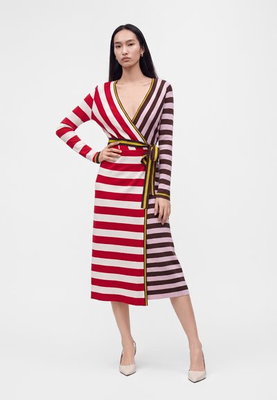 Femme debout, main sur la hanche, portant une robe portefeuille mi-longue à rayures horizontales rouges, blanches, violettes et marron, et des talons pointus beiges.