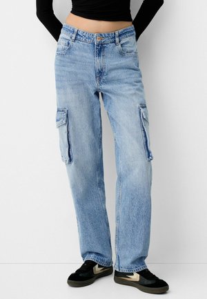 Person trägt hellblaue Denim-Cargohosen mit Seitentaschen, ein schwarzes, langärmeliges, kurzes Oberteil und schwarze Turnschuhe mit beigen Chevron-Details.