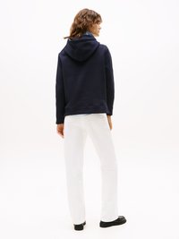 Une femme aux cheveux bouclés se tient de profil à droite, portant un sweat à capuche navy, un pantalon blanc, des chaussures noires et un col de chemise bleu clair apparent.