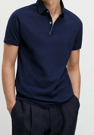 Mann trägt ein dunkelblaues Polohemd mit kurzen Ärmeln, zwei Knöpfe geöffnet, Hände in den Taschen einer dunklen Hose, steht vor einem einfarbigen Hintergrund.