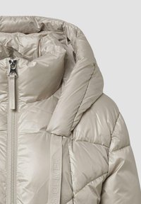 Jasno beżowa kurtka puffer z błyszczącym, pikowanym wykończeniem; posiada wysoki kołnierz, pełny zamek błyskawiczny oraz markowe szczegóły z materiału po boku.