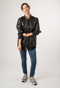 Zwarte leren bomberjack met geribbelde manchetten en zoom, gecombineerd met blauwe jeans en grijze sneakers. Model staat met gekruiste armen.