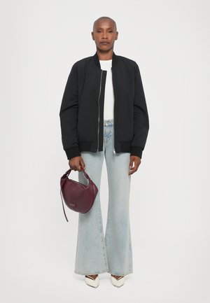 Cappotto bomber nero con polsini a coste, abbinato a jeans a campana blu chiaro. Porta una borsa in pelle bordeaux e indossa tacchi bianchi a punta.