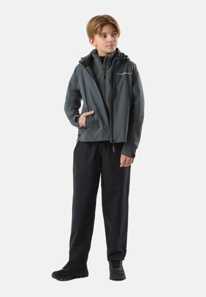 Jongen staand, draagt een donkergrijze capuchonjas over een fleecejas met ritssluiting, zwarte broek en zwarte wandelschoenen, met één hand in de jaszak.