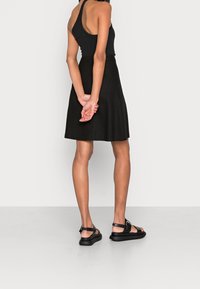 Schwarzes ärmelloses Kleid mit einem ausgestellten Schnitt und strukturiertem Stoff, kombiniert mit flachen schwarzen Sandalen mit einem Schnallenriemen.