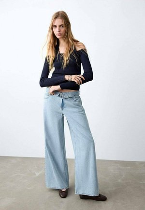 Junge Frau mit langen blonden Haaren, die ein schwarzes, schulterfreies, geripptes Oberteil, hellblaue Jeans mit weitem Bein und dunkelbraune Schuhe trägt, vor einfarbigem Hintergrund.