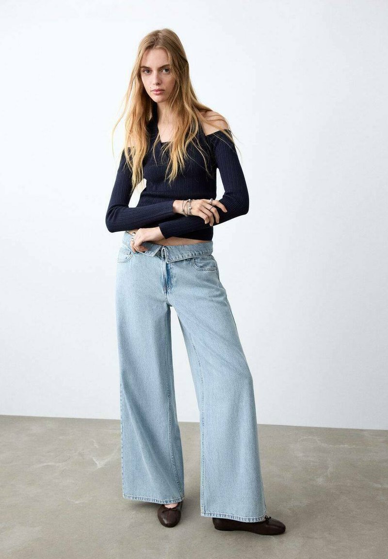 Jeune femme aux longs cheveux blonds portant un haut côtelé noir à épaules dénudées, un jean large bleu clair et des chaussures marron foncé sur un fond uni.