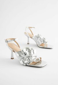 Sølv højhælede sandaler med firkantet tå, der har krøllet metalmateriale og en voluminøs design. Justerbar ankelrem medfølger.