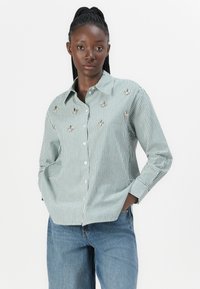 Grön- och vitrandig skjorta med knappar och broderade detaljer, långa ärmar och avslappnad passform, tillsammans med lösa jeans.
