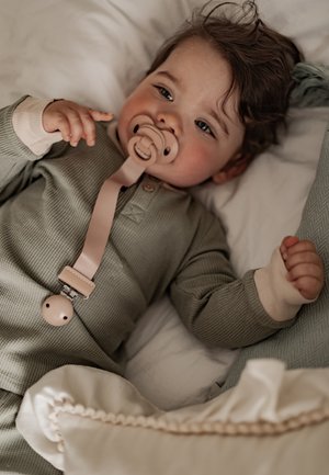 Baby with curly hair lying on a bed, wearing a green onesie and using a beige pacifier attached to a clip on the outfit.