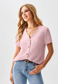 Roze T-shirt met korte mouwen, een golvende halslijn en knopen aan de voorkant, gemaakt van getextureerde stof; gecombineerd met lichtblauwe spijkerbroek en een riem.