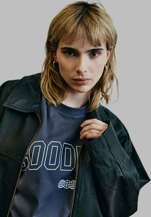 Black leather jacket with a wide collar over a dark blue sweatshirt featuring bold white text. Model has short blonde hair and bold earrings.