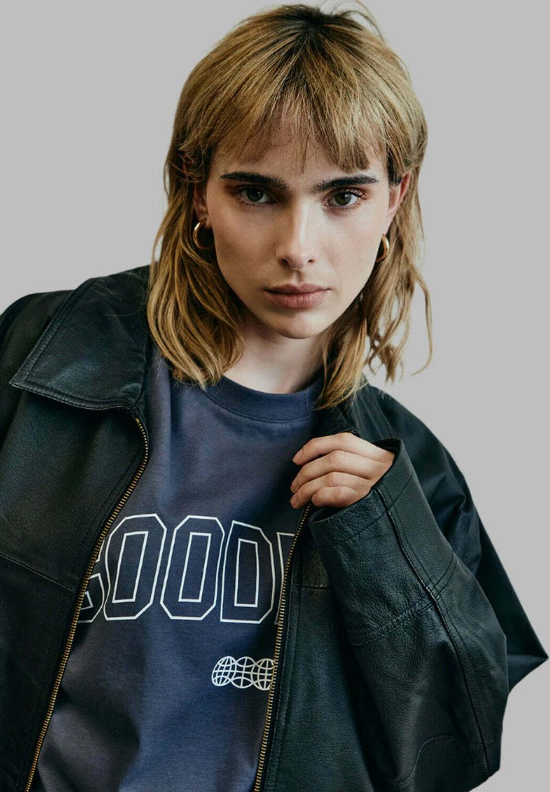 Black leather jacket with a wide collar over a dark blue sweatshirt featuring bold white text. Model has short blonde hair and bold earrings.