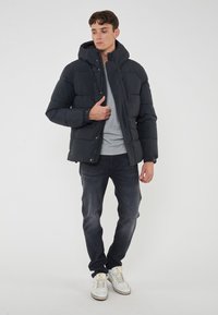 Giubbotto imbottito nero con cappuccio, design trapuntato e zip frontale, indossato sopra una maglietta grigia, abbinato a jeans scuri e sneakers bianche.