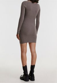 Kvinna iklädd en åtsittande, långärmad, ribbad miniklänning i grått med svarta, kraftiga stövlar till mitten av vaderna, stående mot en enfärgad bakgrund.