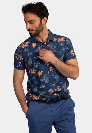 Man die de kraag van een blauw poloshirt met oranje bloemenprint aanpast, draagt blauwe broek en zwarte riem, staat voor een witte achtergrond.