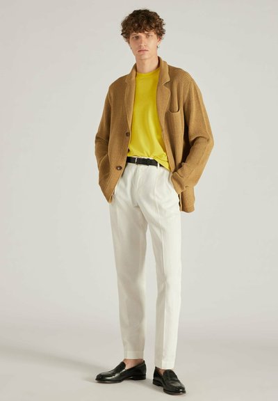 Cárdigan beige de canalé con abertura frontal, combinado con una camiseta amarilla brillante, pantalones blancos y mocasines negros. Diseño simple y texturizado en toda la prenda.