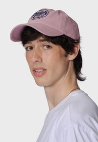 Rosa bomull-basebollkeps med en marinblå broderad logotyp framtill. Har en böjd skärm och justerbart spänne på baksidan.